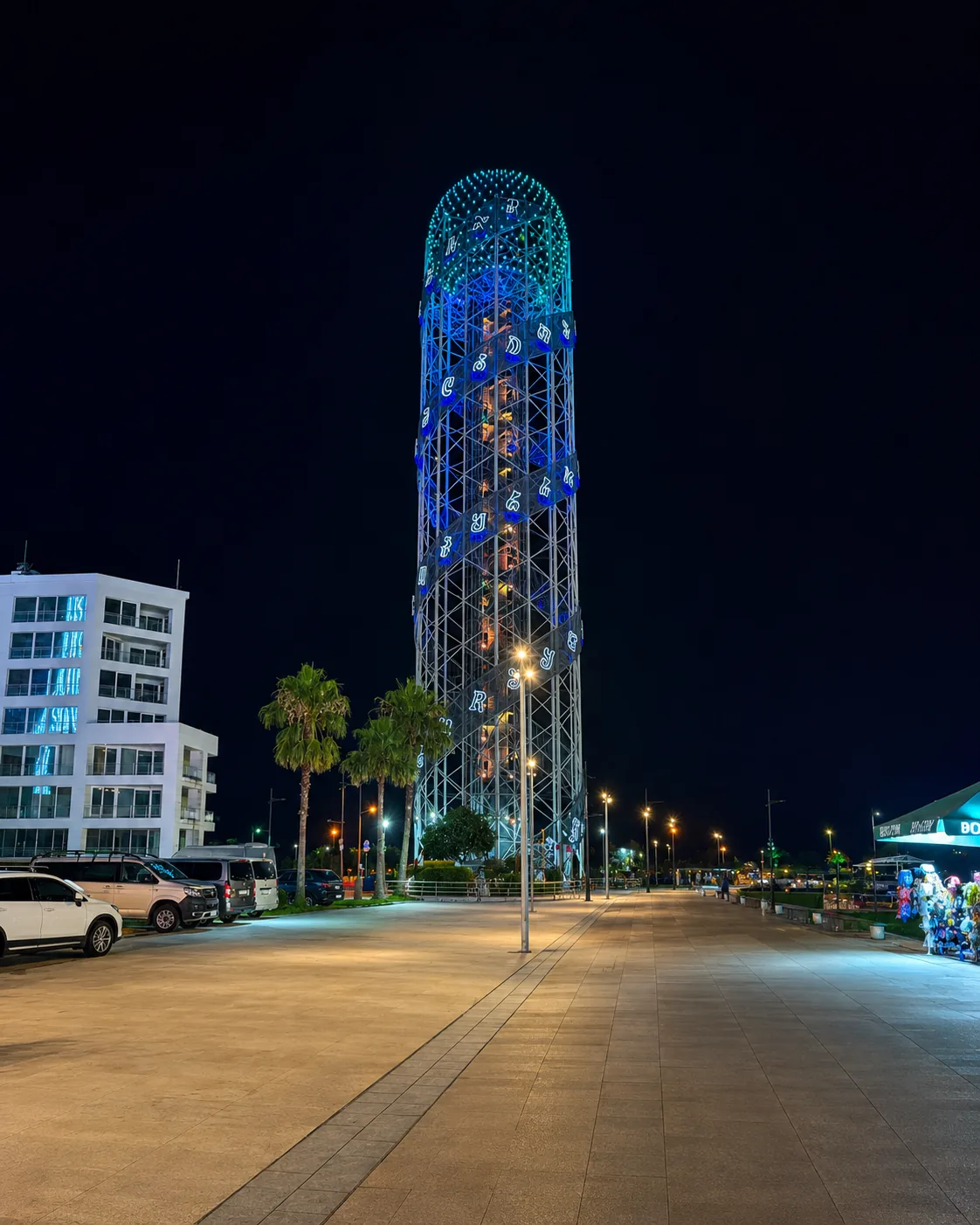 Abecední věž v Batumi zářící v noci modře, při pohledu z promenády bulváru s palmami, Gruzie