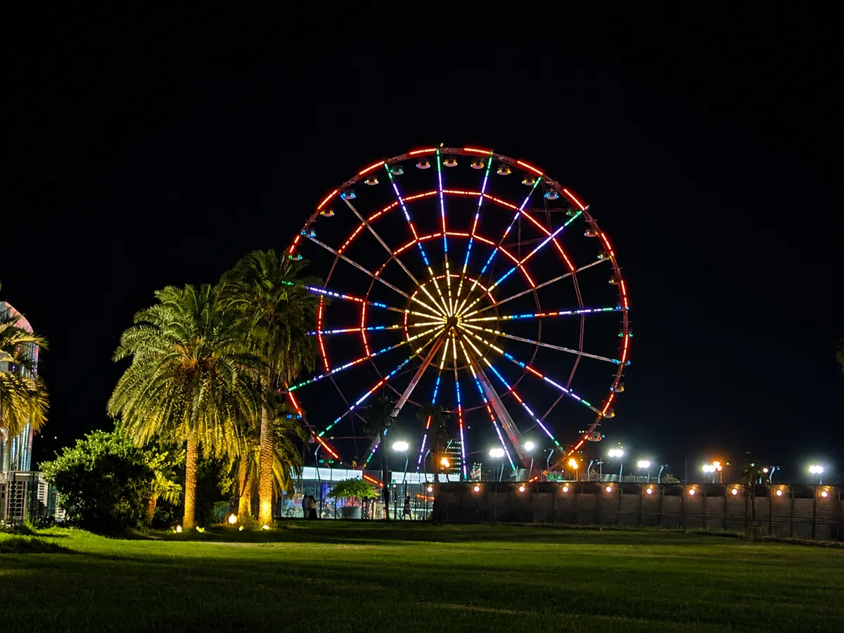 A brightly lit Ferris wheel glowing in rainbow colours at night beside palm trees in Batumi's seaside park, Georgia