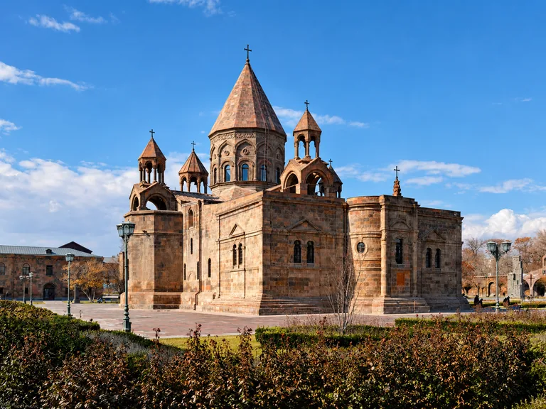 The UNESCO-listed Mother Cathedral of Holy Etchmiadzin — a cruciform tuff-stone church with a central conical dome and corner bell-towers, behind autumn shrubs under a blue sky, Vagharshapat, Armenia