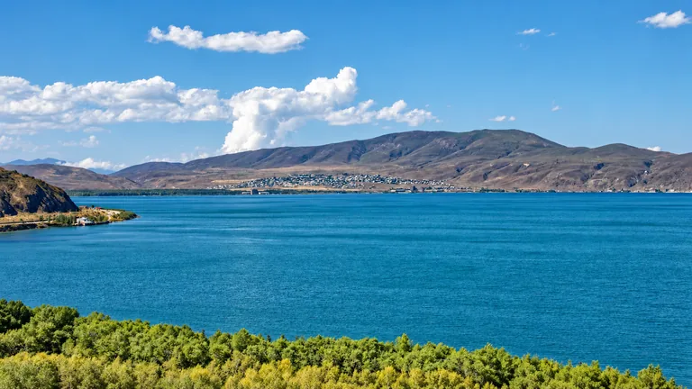 The deep blue water of Lake Sevan under a summer sky, with a wooded shore in the foreground and a lakeside village below bare hills on the far shore, Gegharkunik, Armenia