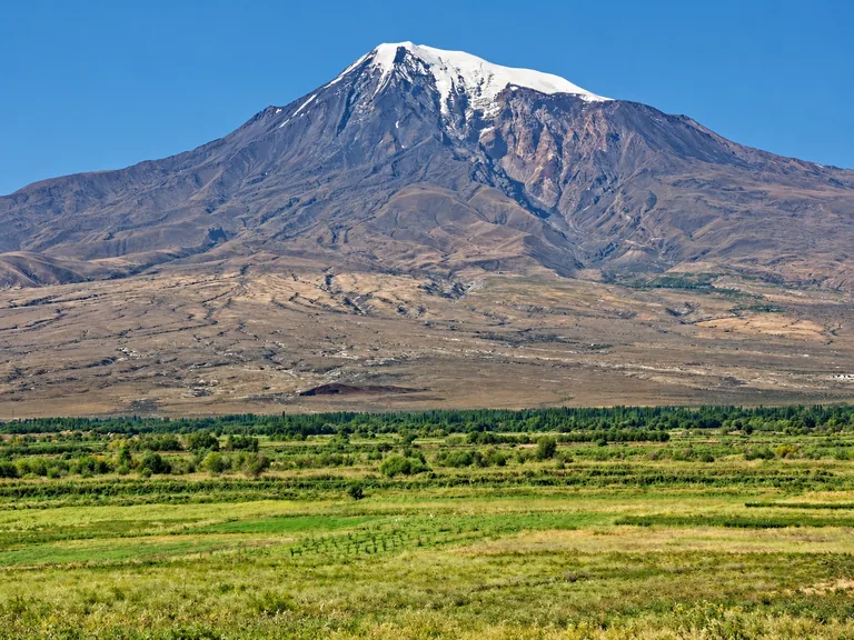 Snow-capped summit of Mount Ararat (Greater Ararat) rising above brown volcanic slopes and the green Ararat plain under a clear blue sky, seen from Armenia — the mountain lies across the border in Turkey