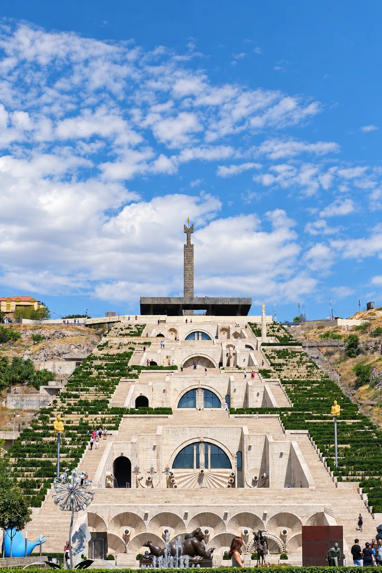 The Yerevan Cascade — a monumental limestone stairway with terraced gardens, fountains and modern bronze sculptures, rising toward a Soviet-era obelisk under a blue sky, Yerevan, Armenia